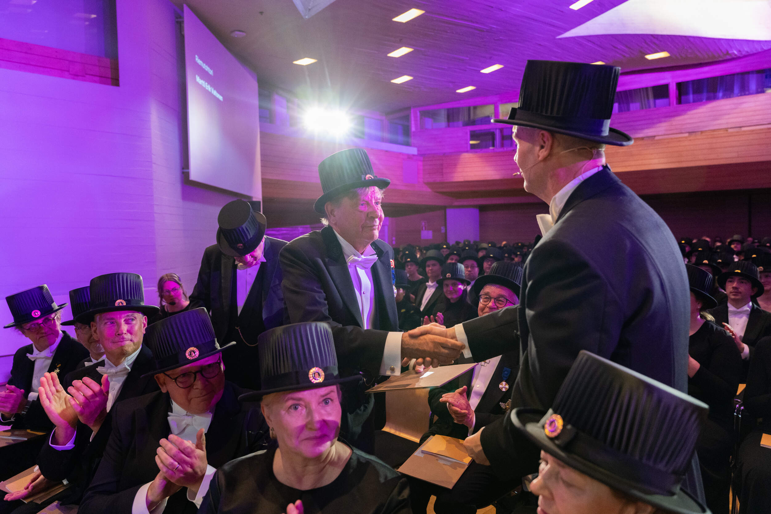 Formal ceremony in purple-lit hall, two people in black tailcoats and top hats shaking hands