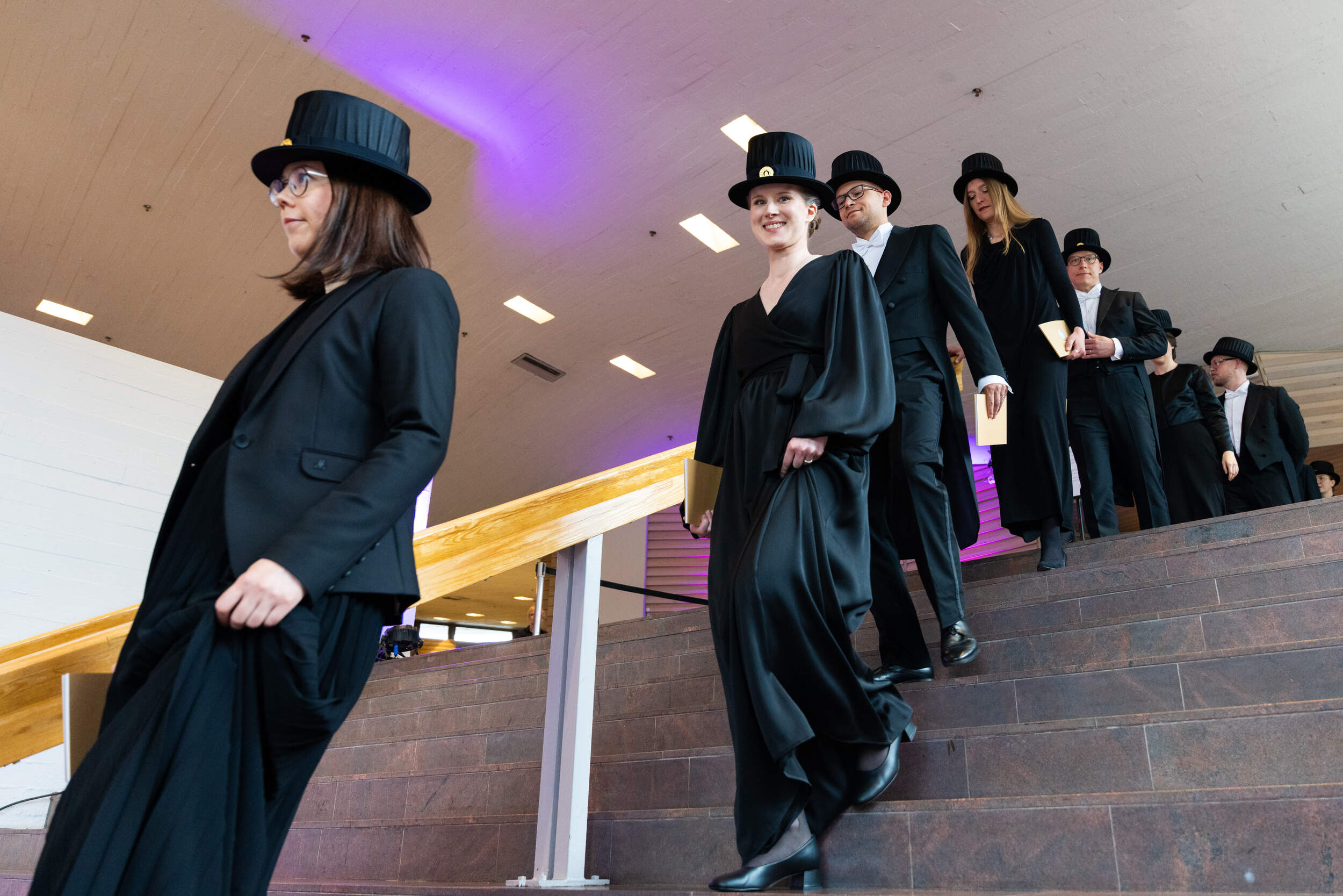 People in black formal dress and top hats walk down indoor stairs holding light-coloured books