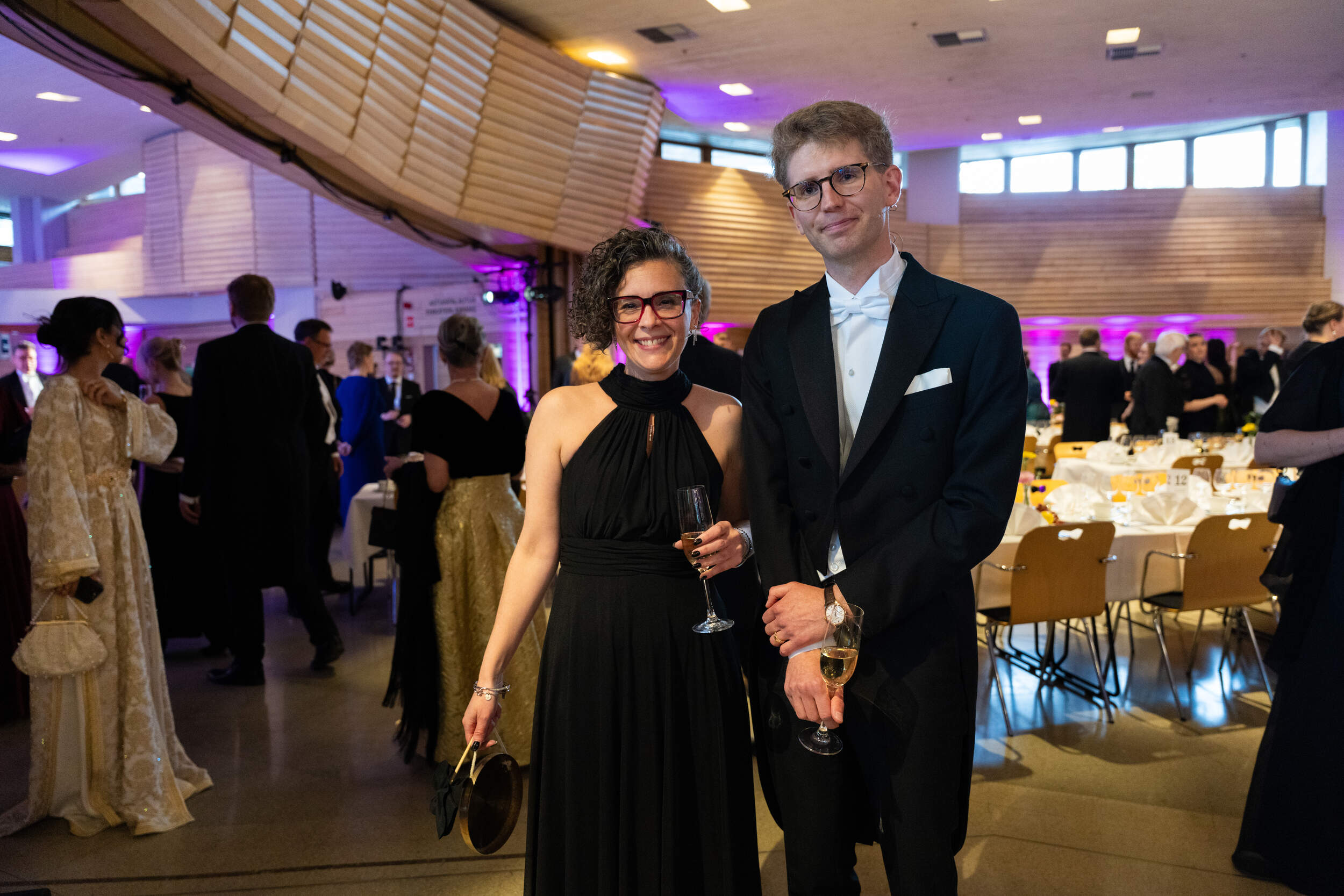 Well-dressed couple holding champagne glasses at a formal indoor gala with round tables and guests in evening wear
