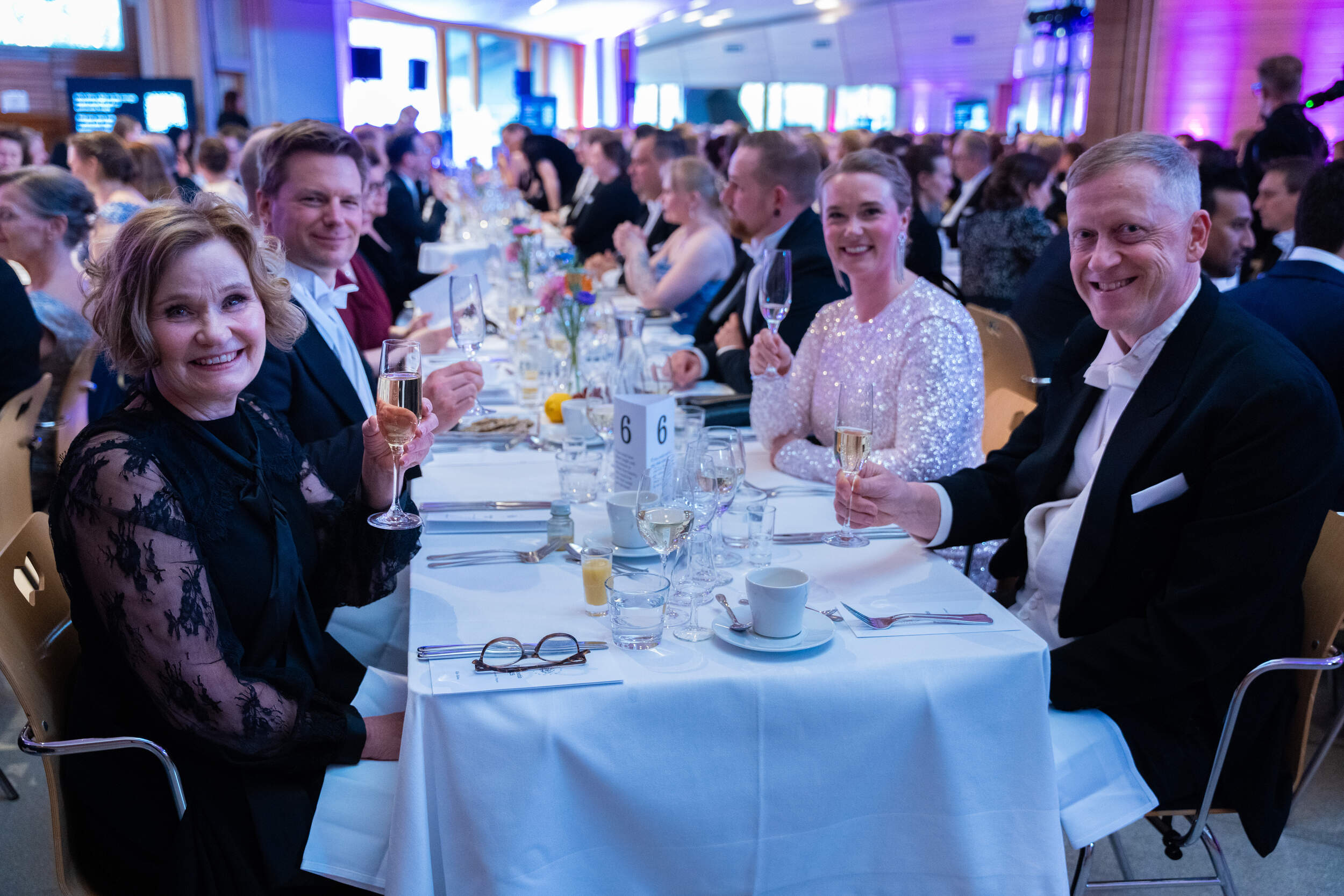 Guests in formal wear sit at a long white‑clothed banquet table, holding champagne flutes