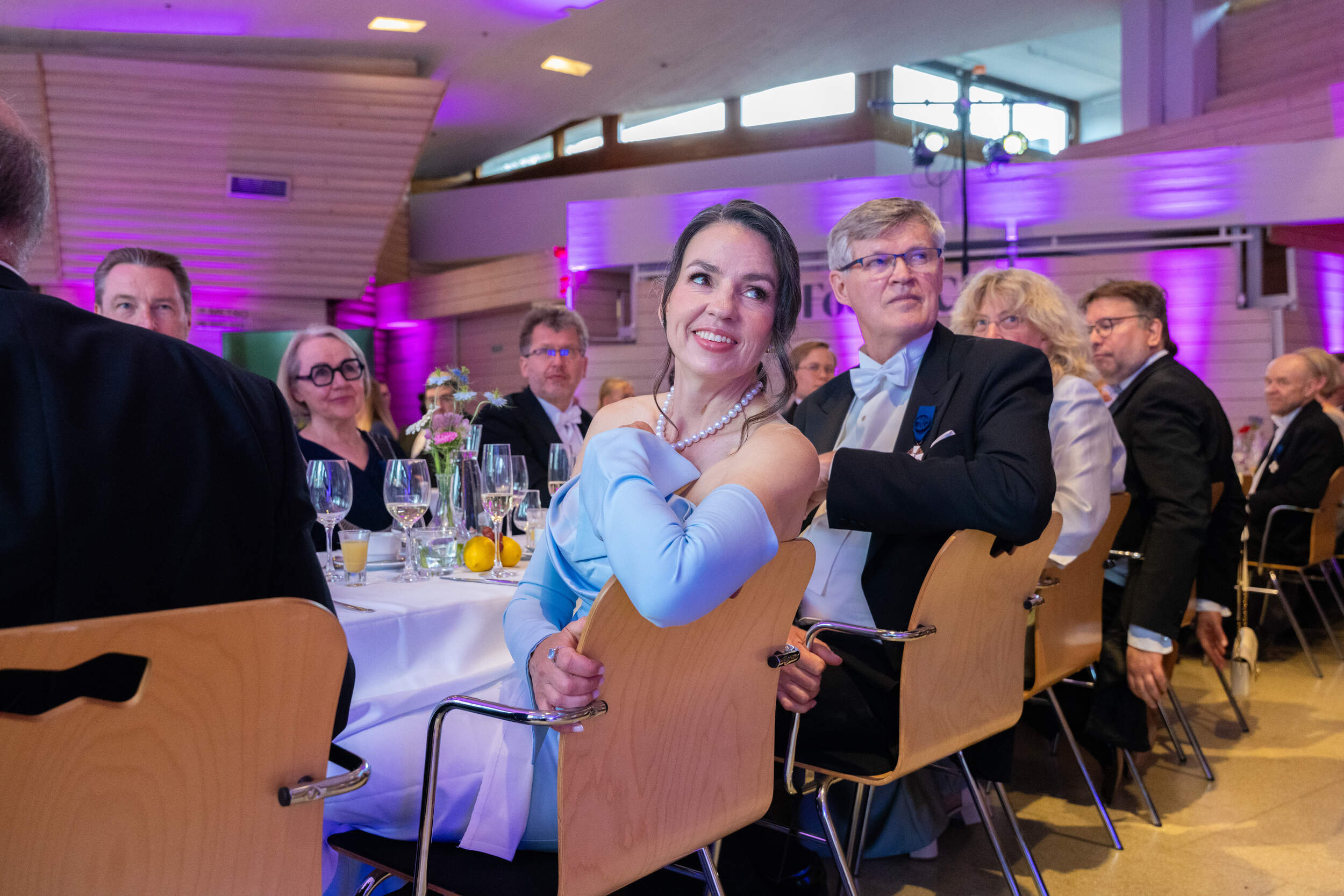 Formal dinner guests seated at round tables with drinks in a hall lit by pink and purple lights