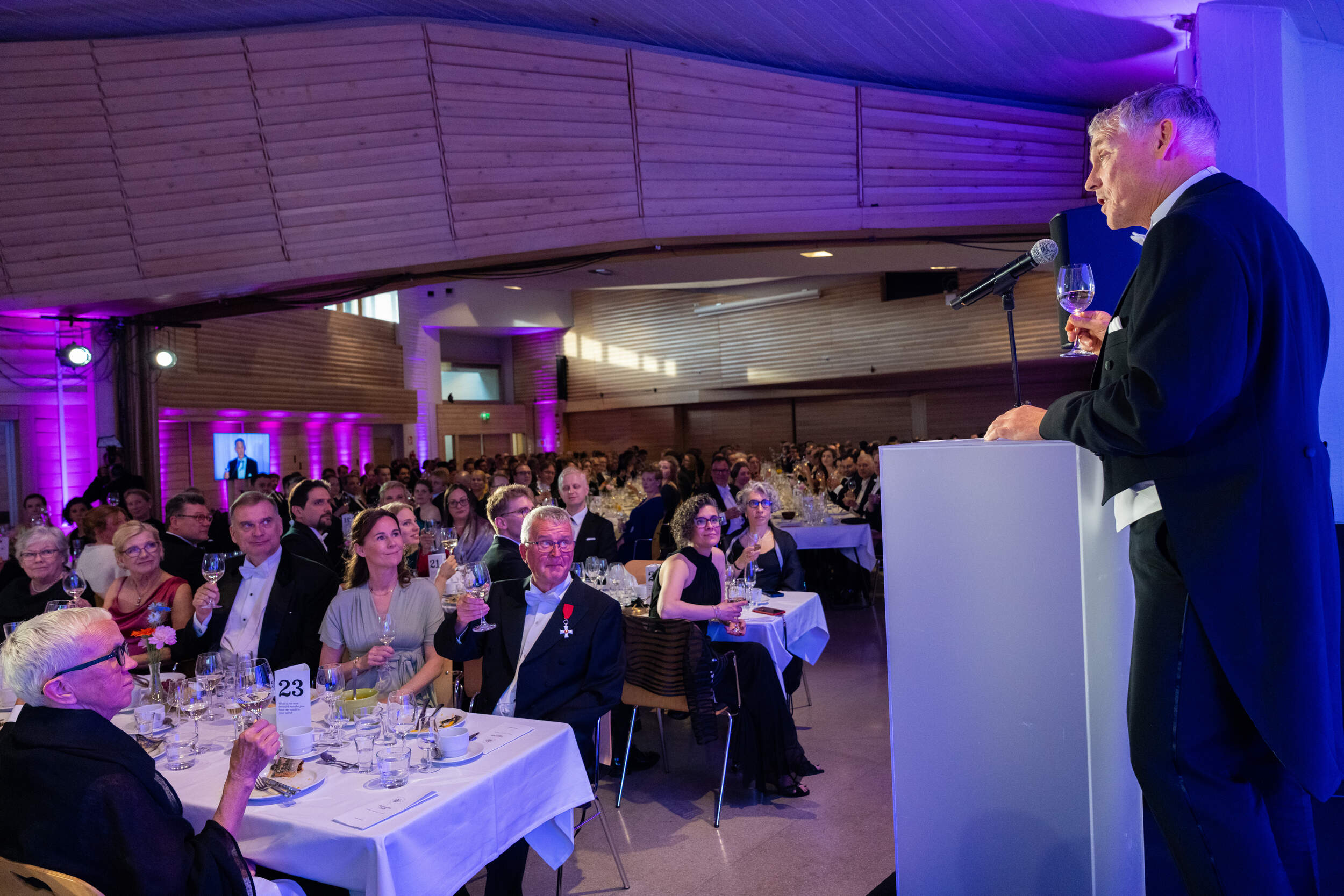Speaker in black tailcoat giving a toast to a seated banquet audience in a purple-lit wooden hall