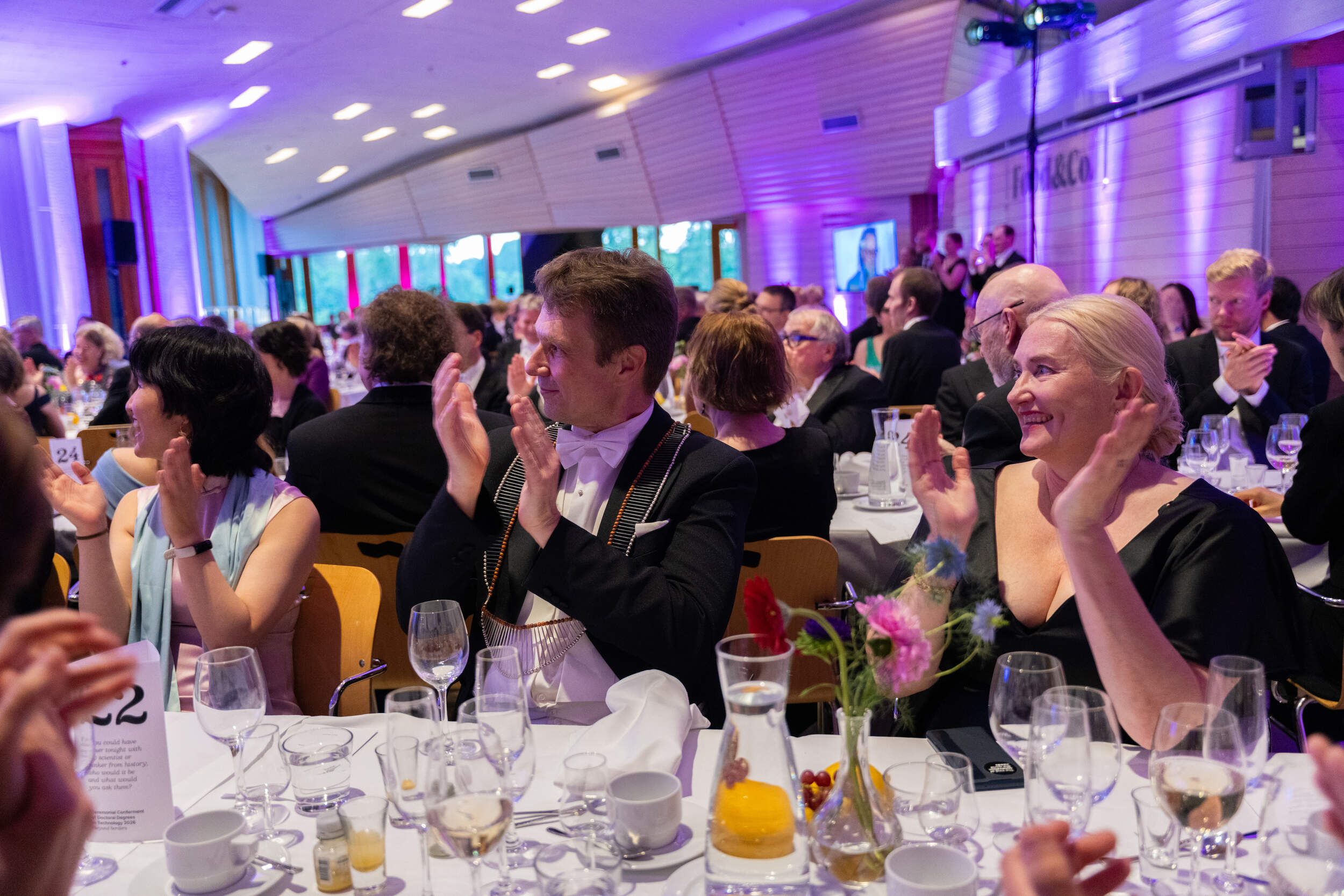 Guests in formal wear sit at round tables, clapping during an indoor gala dinner with purple lighting.