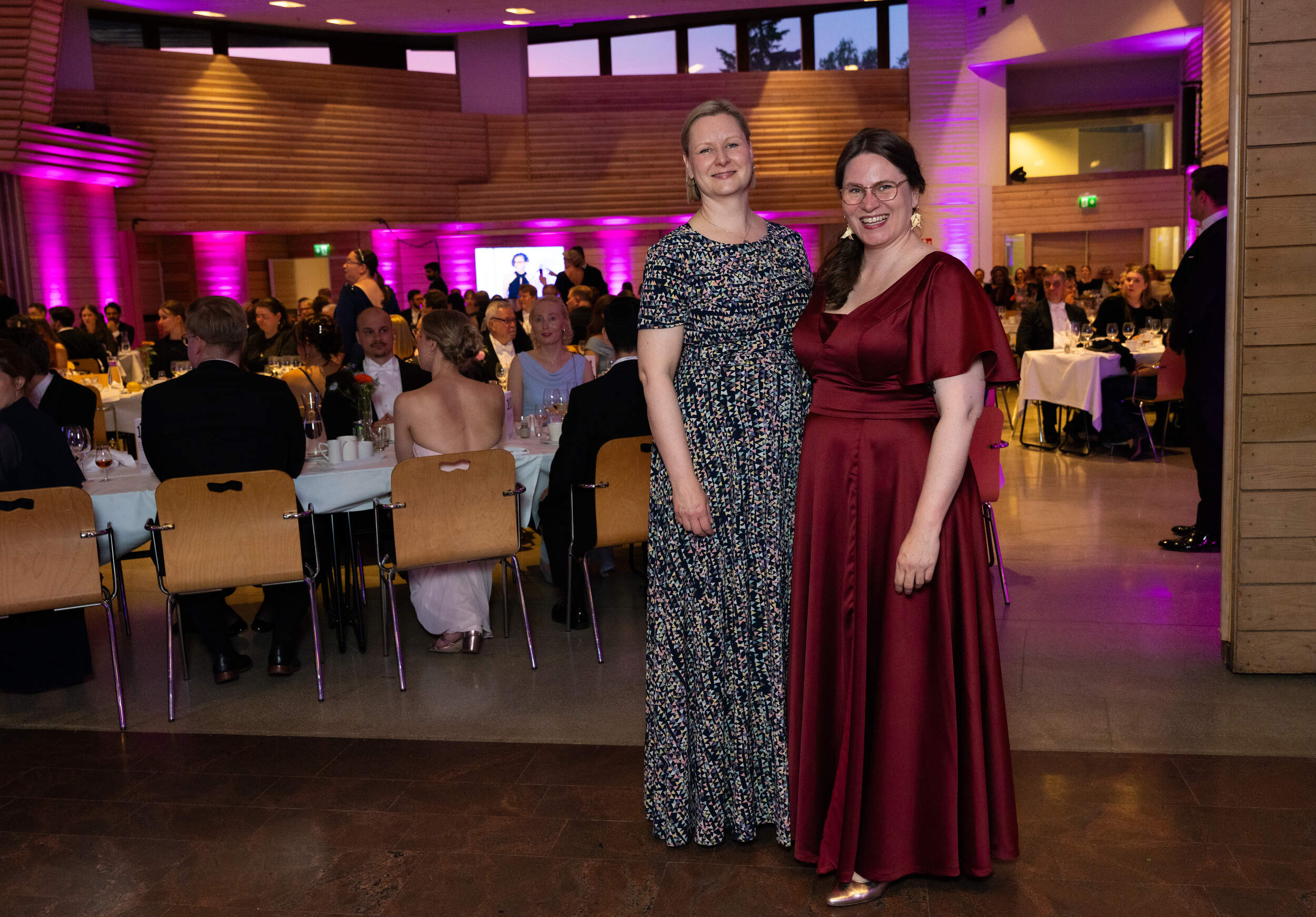 Two women in long dresses pose at a formal dinner, guests seated at tables in a purple-lit hall