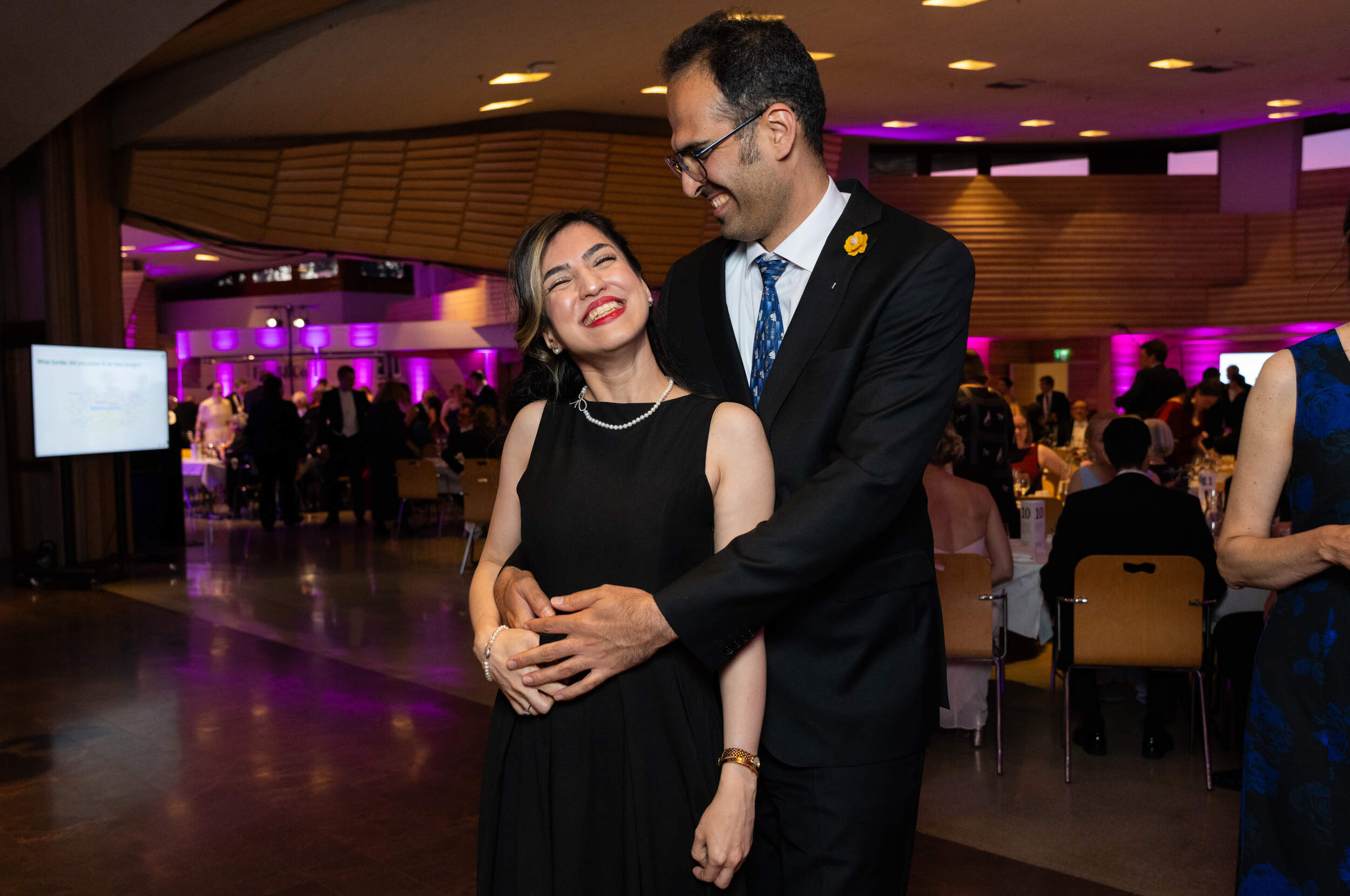 Couple in formal black outfits embrace on a ballroom floor, with seated guests and purple lights behind them.