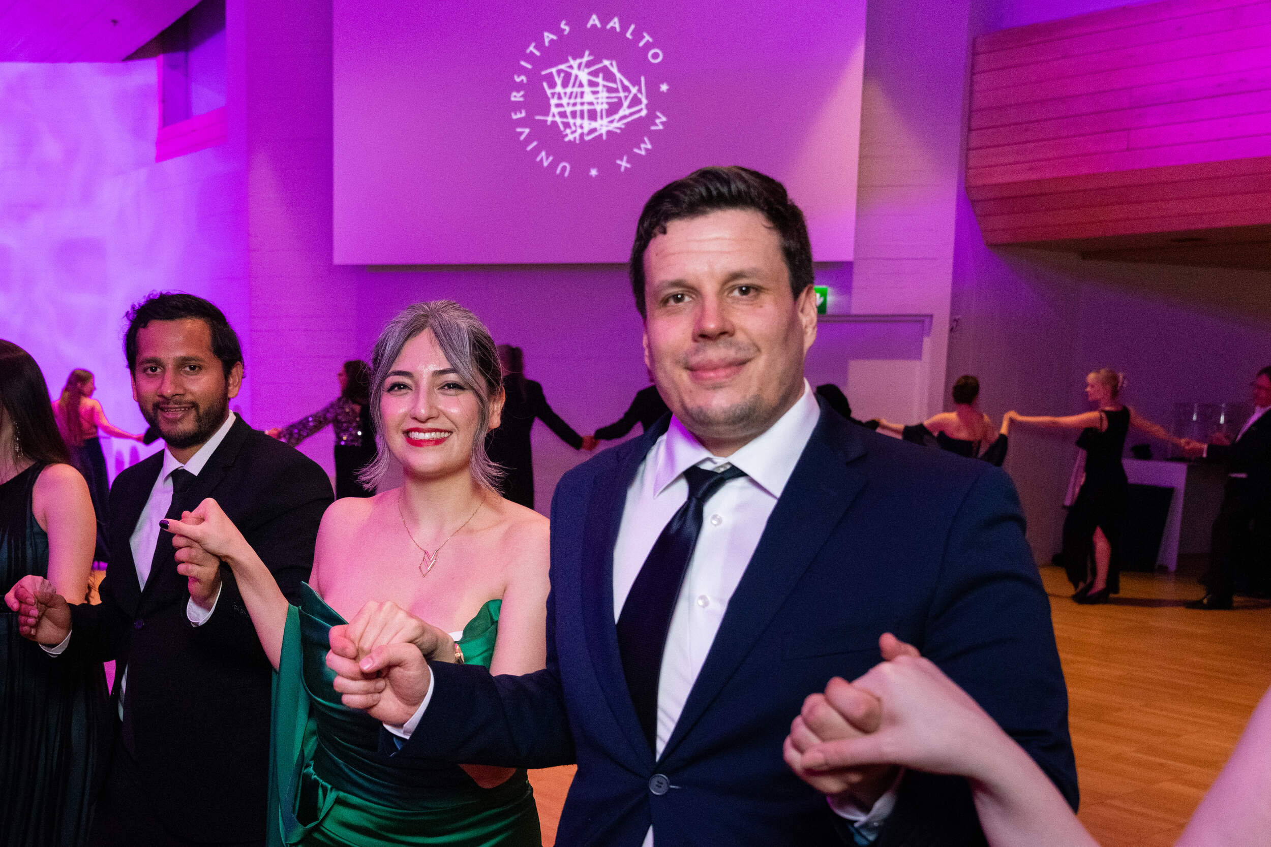People in formal dress dance in a ballroom with purple lighting and an Aalto University logo on the wall.
