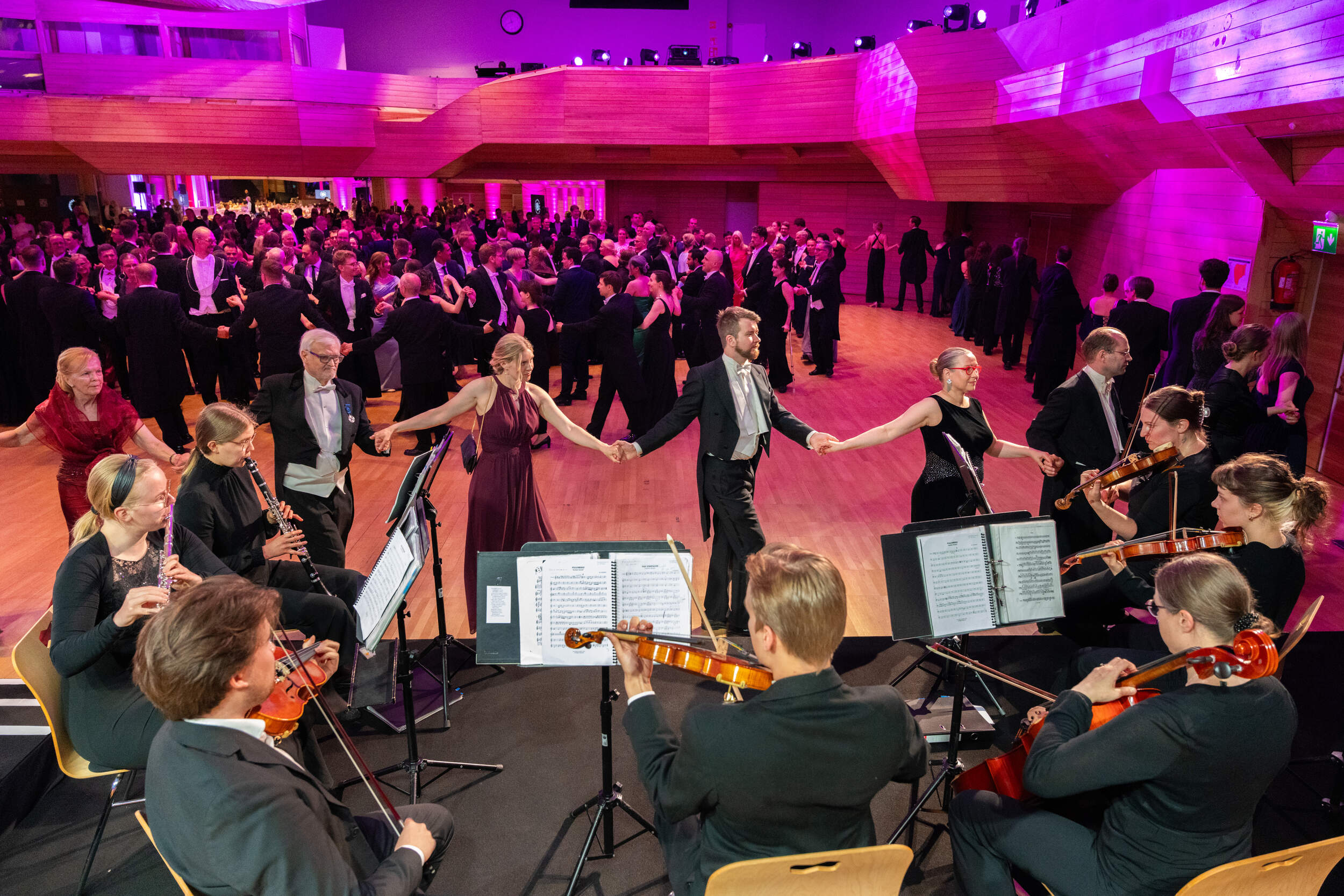 Orchestra plays on stage while couples in formal dress waltz in a bright pink-lit ballroom