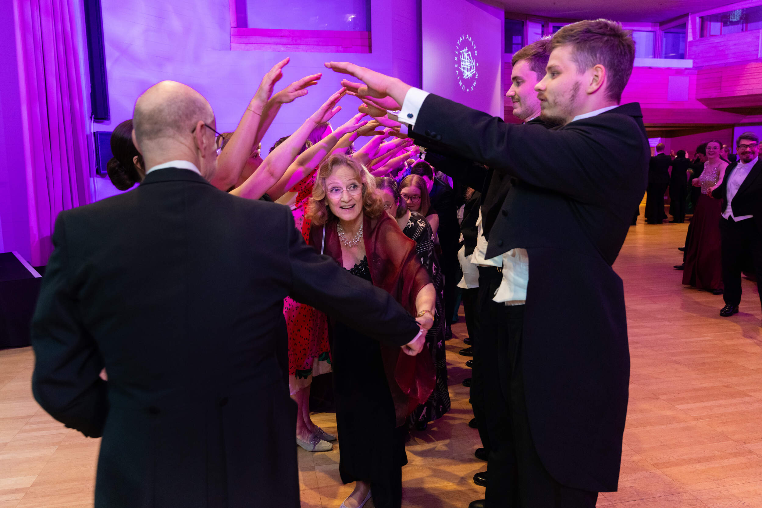 People in formal wear form an arm tunnel for dancers at a purple-lit ballroom event