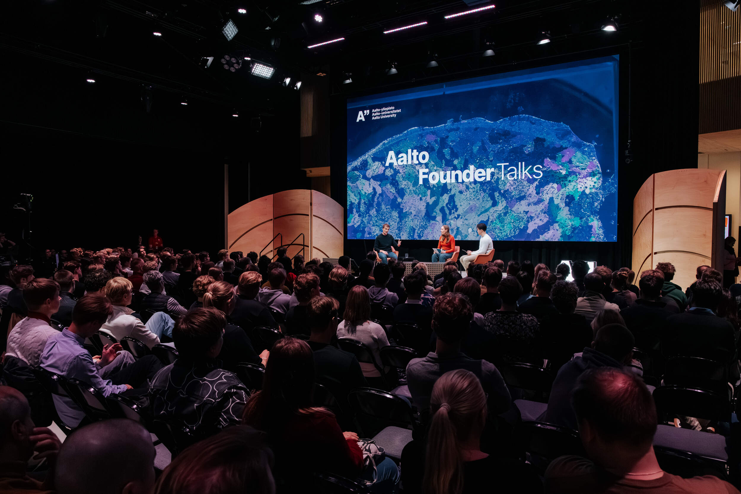 Yleisö seuraa kolmen puhujan Aalto Founder Talks -lavaesitystä hämärässä salissa suuren sinisen näytön edessä.