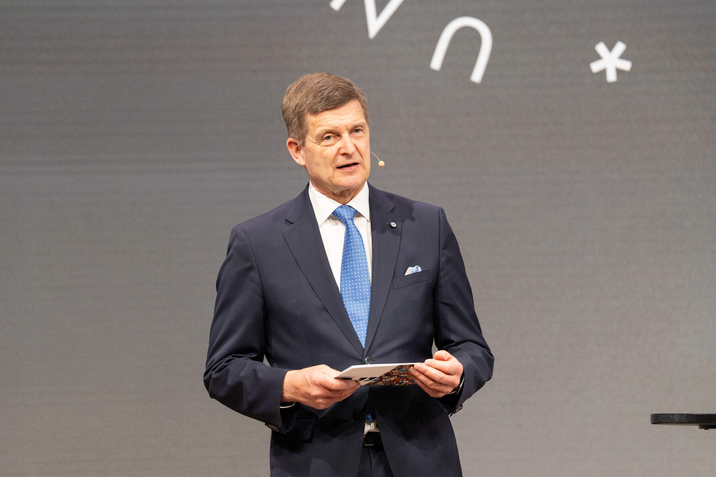 President Ilkka Niemelä in dark suit and blue tie speaks on stage, holding notes, with symbols on grey backdrop.