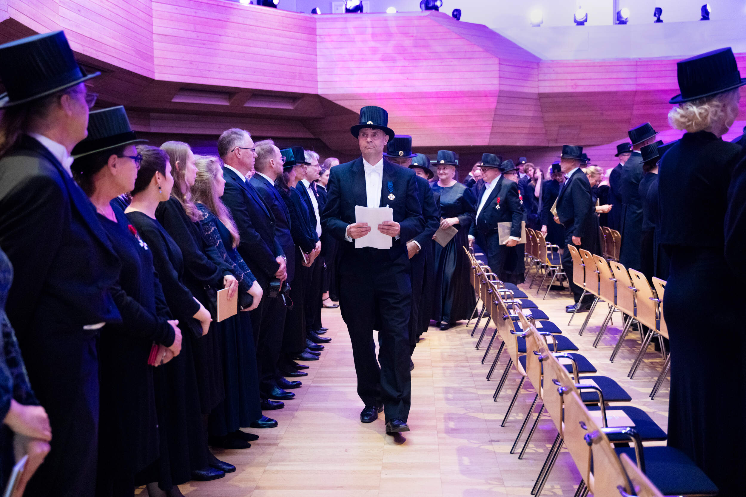 Formal ceremony with people in black tailcoats and top hats walking in a wooden hall