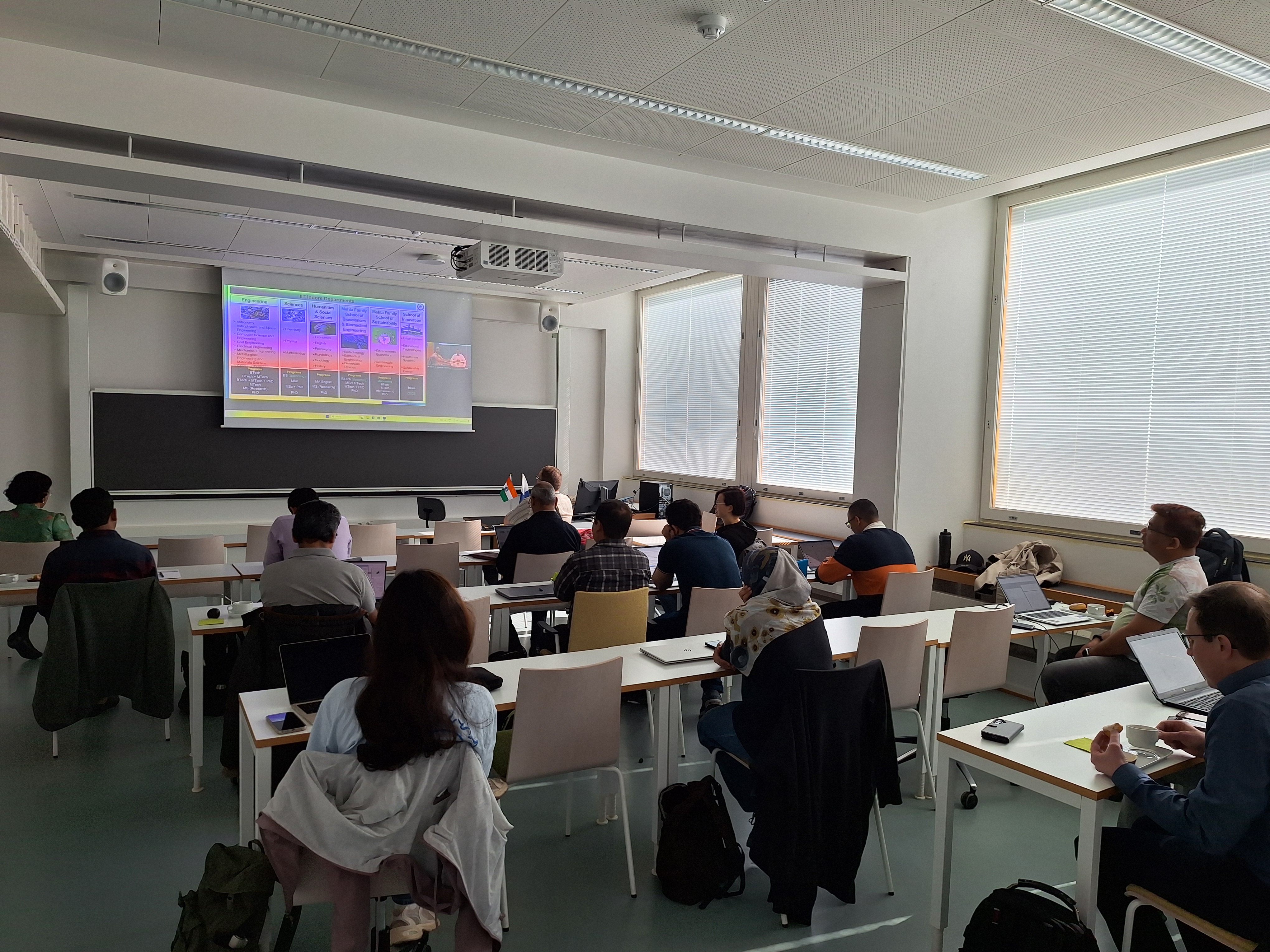 Students sit at desks with laptops in a bright classroom, watching a colourful slide on a projector screen.