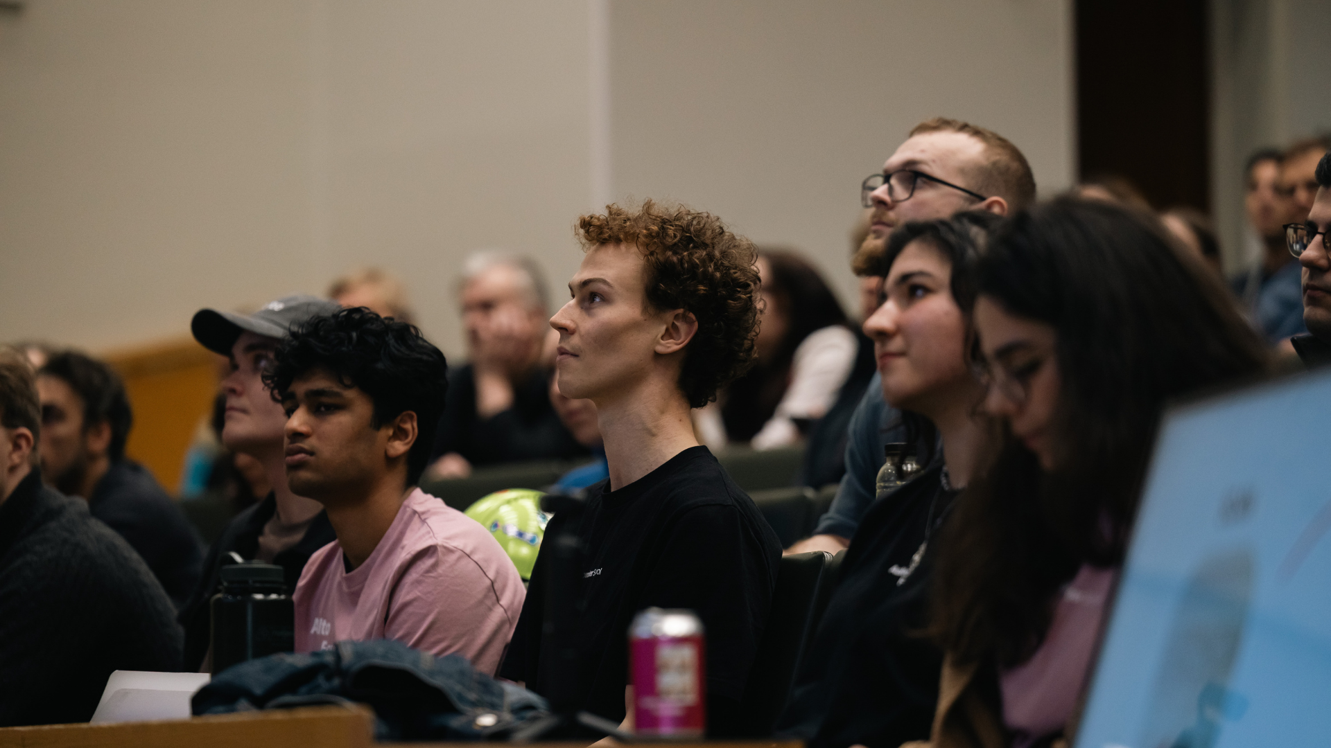 Students with blurred faces sit in a lecture hall, listening, with laptops and a pink drink can visible.