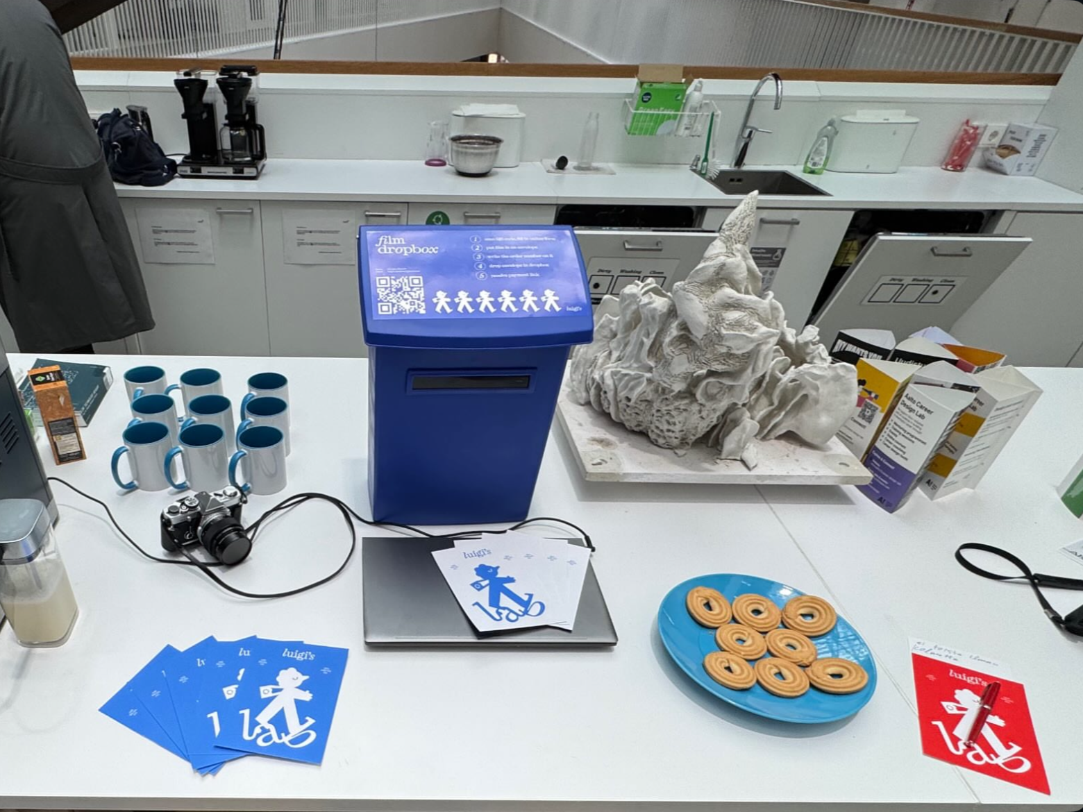 Office kitchen counter with blue film dropbox, camera, mugs, cookies on a plate and a white sculpture.