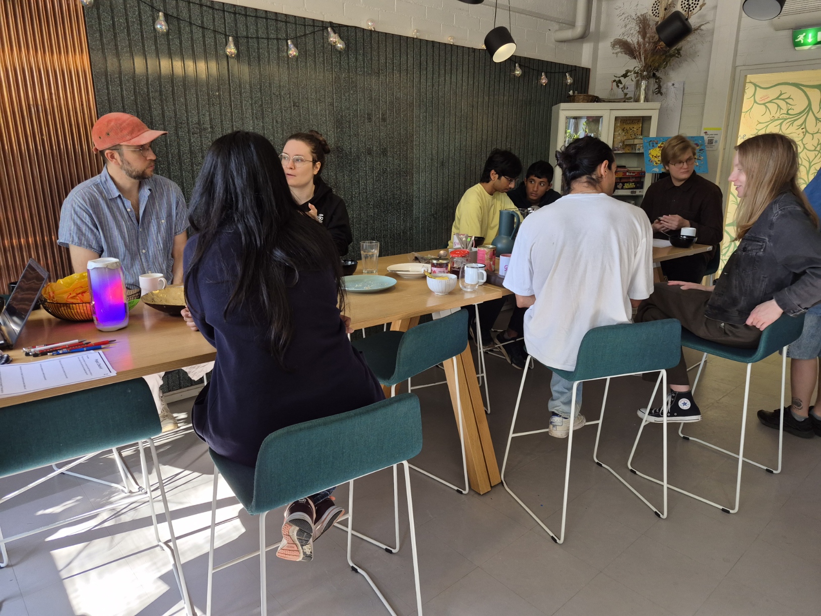 Group of people chatting around a long wooden table with drinks and snacks in a bright kitchen