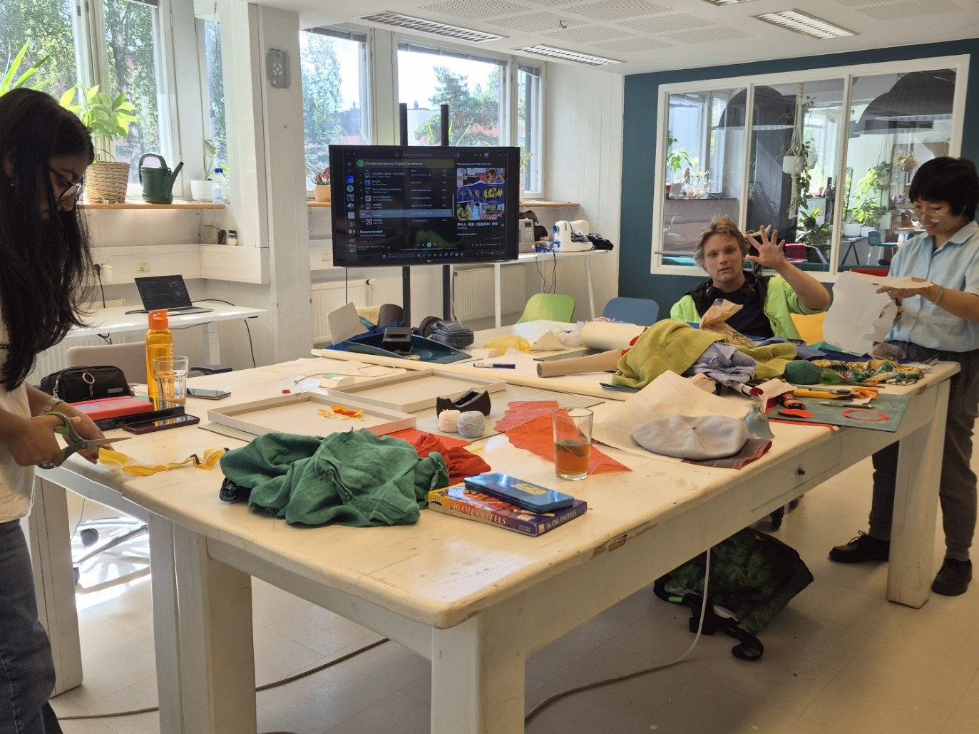 Three people crafting at a large white table covered with colourful fabrics and art supplies in a bright studio