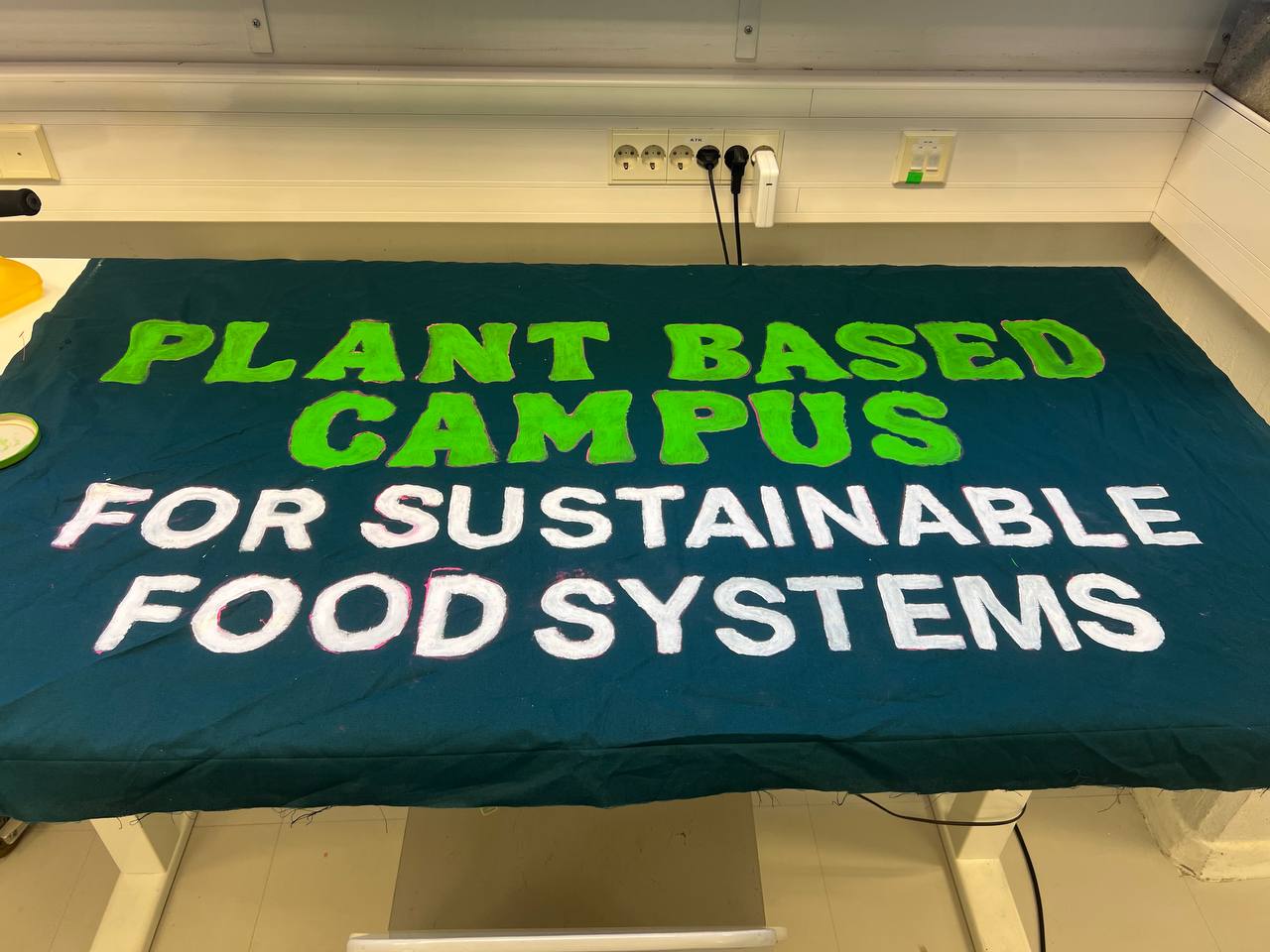 Hand-painted green and white banner on a table reading: PLANT BASED CAMPUS FOR SUSTAINABLE FOOD SYSTEMS