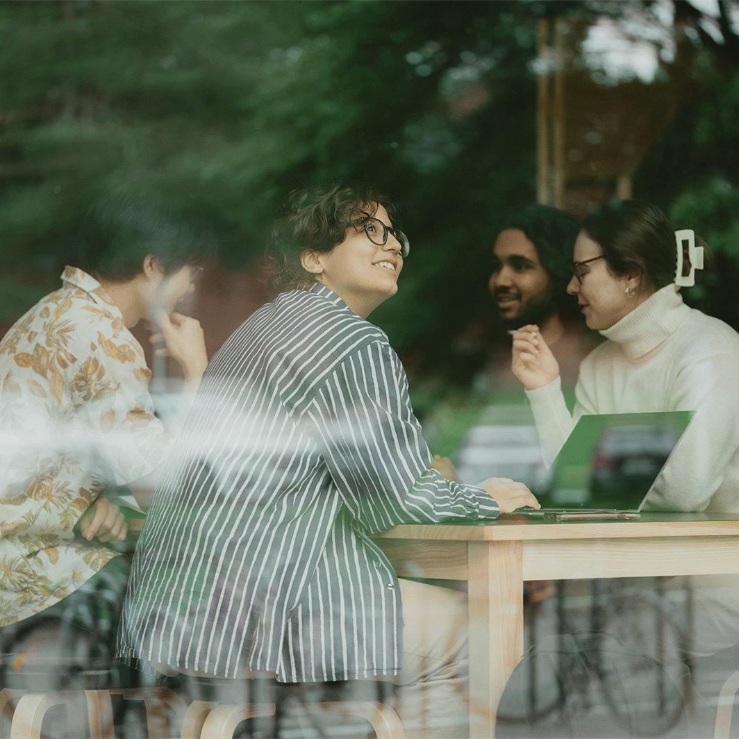 Three people sit at a wooden table with a laptop, viewed through a window with reflections of greenery and parked cars.