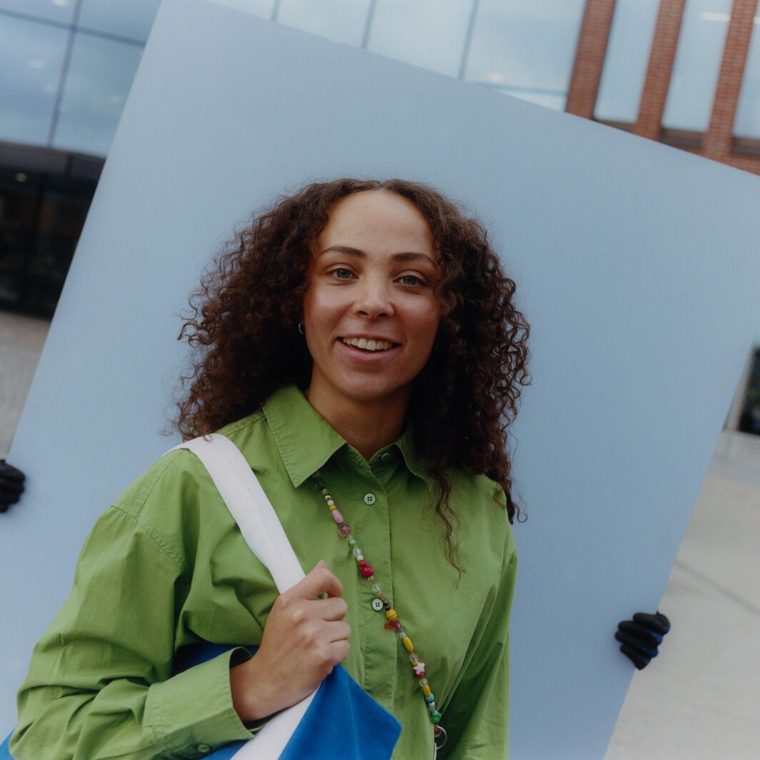 Person in green shirt holding blue bag stands before large grey board held by gloved hands outdoors