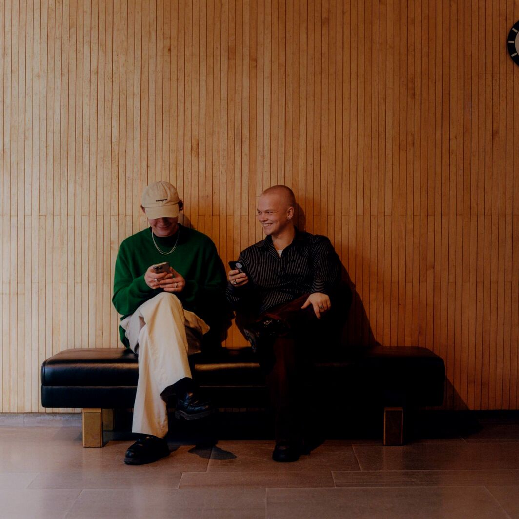 Two people on a black bench using phones, against a wooden wall with a round clock above them