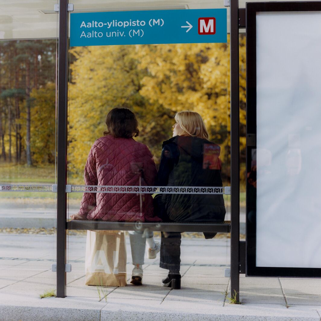 Two people sit at a glass bus stop near Aalto University, autumn trees with yellow leaves behind them