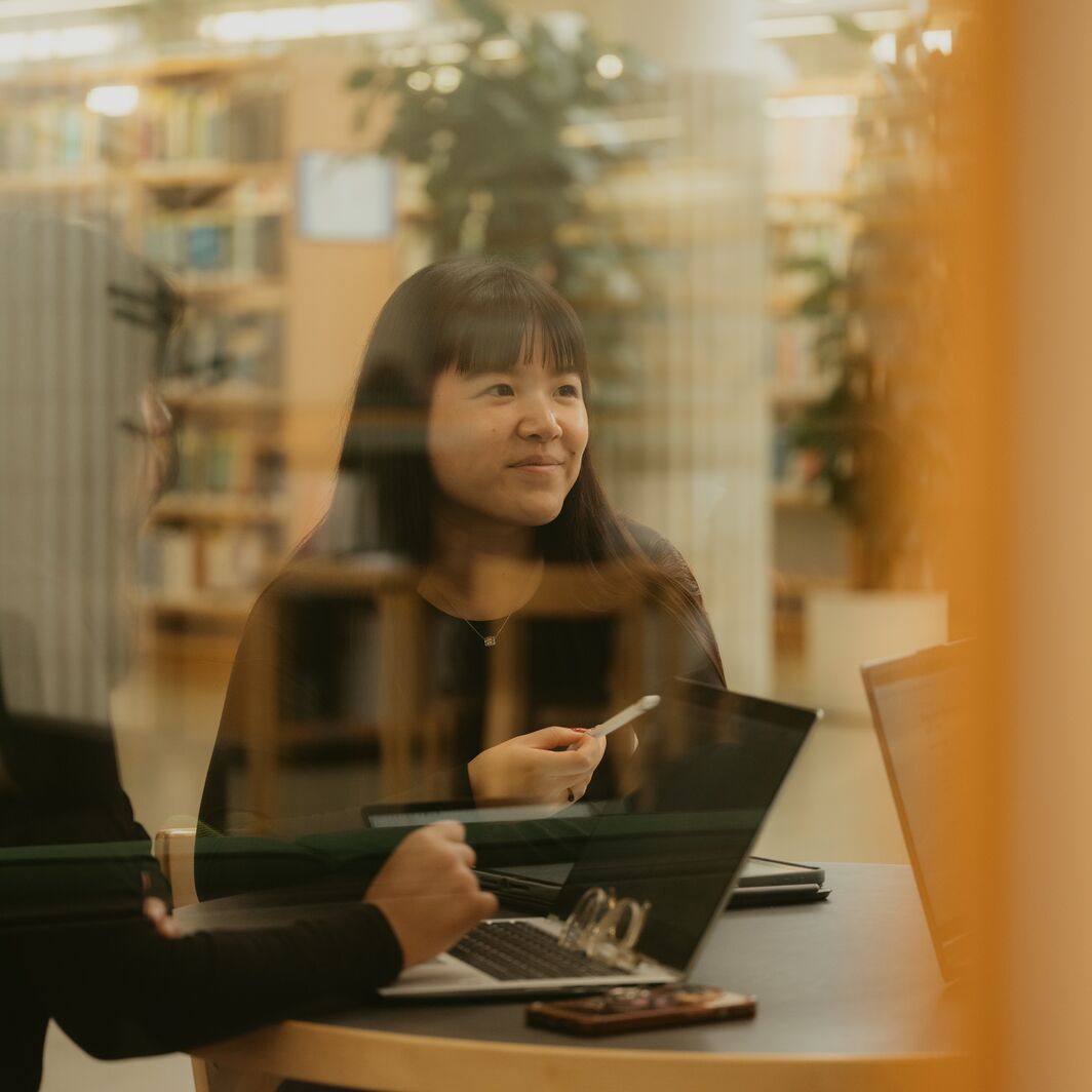 Woman with long dark hair in a library meeting room, holding a pen by laptops, seen through glass.