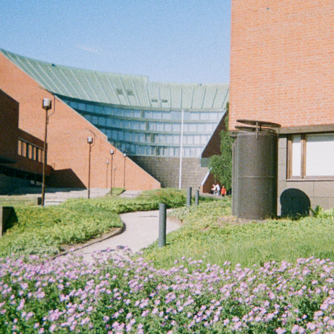 Red brick campus buildings with glass roof and flower beds along a curving path in sunlight