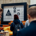 three men talking to each other in the foreground, while another person with back turned looks at the A Grid sign on the background