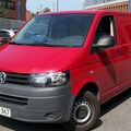 Punainen Volkswagen-pakettiauto on pysäköitynä parkkipaikalle, taustalla muita autoja ja rakennuksia.