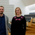A man and woman in casual wear, dark tones, smiling at the camera inside the Aalto Hall.