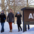 Three people walking in winter next to a sign that says 'Aalto University' with snow-covered trees and buildings in the background.