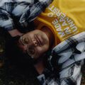 Person lying on grass wearing a yellow shirt with white text and a black-and-white checkered jacket.