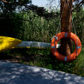 A yellow kayak and an orange life buoy are propped against trees by a lakeside, with tall grass and reeds in the background.
