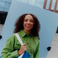 Curly-haired person in a green shirt holding a colourful lanyard in front of a modern building.