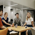 A group of five people sit around a table with mugs, having a conversation in a modern room with white walls.