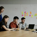 Four people collaborate at a table with laptops. Sticky notes are on a whiteboard behind them, showing various colours.