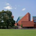 Modern red brick building with glass windows next to trees and a lawn, under a blue sky.