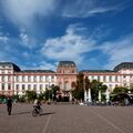 A large, historic building with a pink facade and grey roof in a square with people walking and cycling.