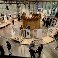 People at an art exhibition with wooden structures displaying artworks. A circular installation made of tall reeds is in the centre.