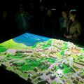 A group of people stand around a lit 3D model of a landscape with buildings and greenery. The model displays 'THE VOICE OF THE NATURE'.