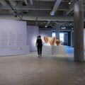 A modern art gallery with pottery, hanging art, and a projection. A person walks past a lavender wall with text.