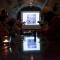 People in helmets and vests sit in a cave-like room, watching a presentation on a screen. A person points at the screen.