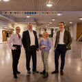 Four people standing in a spacious indoor area with signs above them. The floor is tiled and walls are white.