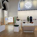 Modern exhibition room with mannequins in green and white clothes, bowls and a beige bathtub on white plinths