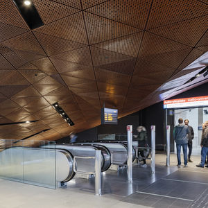 Ihmisiä seisomassa tasanteella, josta johtaa rullaportaat alaspäin metroasemalle. 