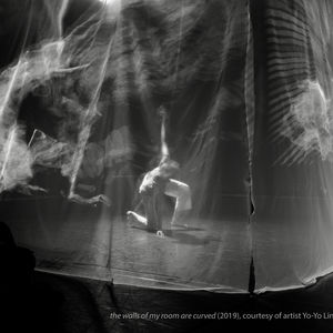 Image Description: black and white photograph of artist Yo-Yo Lin performing on a stage with right knee and her left hand touching the floor. She is surrounded by a translucent scrim with graphical images projected upon it.   the image credits:  the walls of my room are curved (2019). Courtesy of artist Yo-Yo Lin. Photo by Steve Dabal.