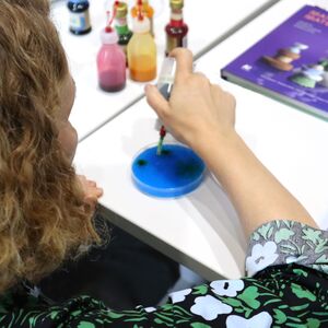 Nainen tiputtelee elintarvikeväriä tippapullosta petrimaljalle selkä katsojaan päin. Taustalla elintarvikevärejä ja Mainiot materiaalit kirja