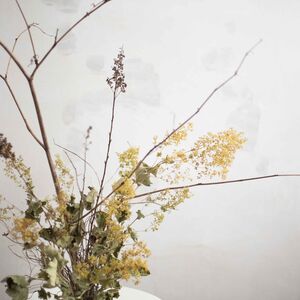 Decorative arrangement of dried branches, yellow flowers, and leaves on a small round white table against a plain wall.