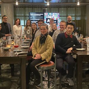 A group of people seated at a long table in a restaurant, drinking and dining, with a presentation screen in the background.