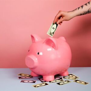 Tatuoitu käsi asettaa setelin vaaleanpunaiseen säästöpossuun. Useita seteleitä sen ympärillä.